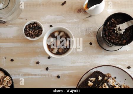 Blick von oben auf Eiskaffee in einer Tasse mit Eiswürfeln, umgeben von Kaffeebohnen, einer Handmahlmaschine, einer kleinen Schüssel mit Bohnen, einem Sirupkrug und Walnüssen auf einem Stockfoto