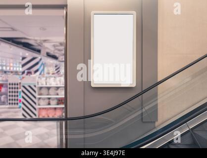 Leeres vertikales Werbeschild in einem modernen Einkaufszentrum neben Rolltreppen mit einem Einzelhandelsgeschäft im Hintergrund, das Platz für Werbeaktionen bietet Stockfoto