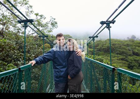 Ein Paar umarmt eine Hängebrücke mit Blick auf die üppige tropische Vegetation des Monteverde Regenwaldes in Costa Rica, umgeben von lebendigen Gr Stockfoto