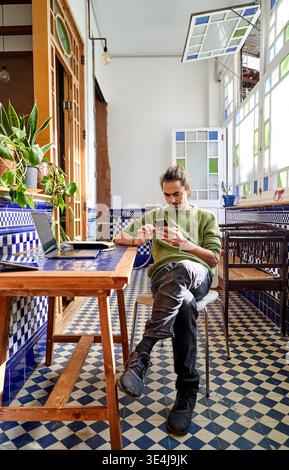 Ein junger Mann sitzt an einem Holztisch in einem hellen Raum. Er arbeitet an einem Smartphone, während ein Laptop auf dem Tisch liegt. Pflanzen sind in der Nähe und verleihen Farbe Stockfoto
