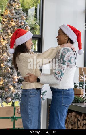 Verschiedene weibliche Partner halten sich in der Hand, tragen Santa-Hüte im Wohnzimmer neben Baum. Paare, festlich, Zweisamkeit, Zuneigung, gemütlich, Wärme, Geschenke || Model veröffentlicht Stockfoto