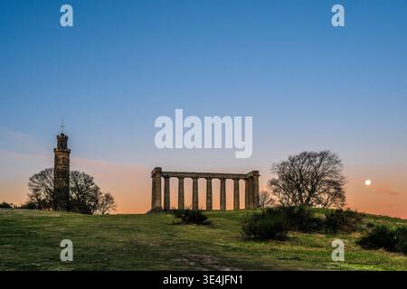 Calton Hill in Edinburgh bei Sonnenuntergang, der das mit Säulen versehene National Monument und den Nelson Monument Tower vor einem pastellfarbenen Himmel mit dem Mond und zeigt Stockfoto