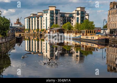 Leith Uferszene zeigt umfunktionierte Binnenschiffe und vertäute Boote neben modernen Wohngebäuden, deren Reflexionen im ruhigen Kanalwasser mit se Stockfoto