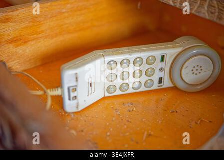 Ein altmodisches weißes Festnetztelefon, das mit Staub bedeckt ist und auf einer Holzoberfläche liegt und numerische Tasten und eine Spiralkabelverbindung zeigt. Stockfoto