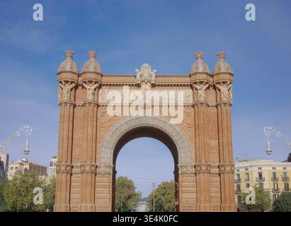 Der kunstvoll verzierte rote Backsteinbogen Arc de Triomf mit Steinfriesen, dekorativen Türmen und Wappen unter einem klaren blauen Himmel in Barcelona, Katalonien Stockfoto