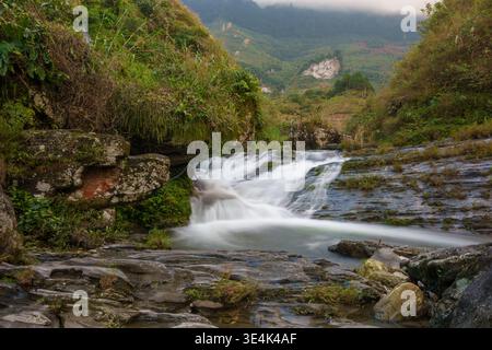Du Gia, Provinz Ha Giang, Vietnam – 18. Januar 2024: Wasser fließt über einen niedrigen Felsvorsprung in ein Bachbecken am du Gia Wasserfall in einem grünen Tal. Stockfoto