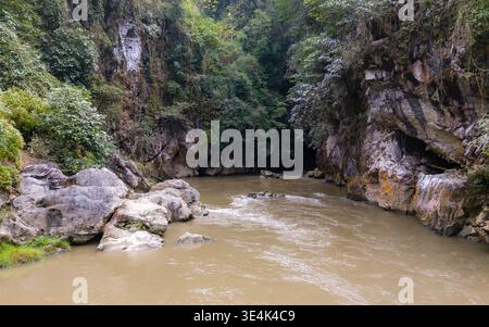 Hang Tham Lin, Vietnam – 18. Januar 2024: Ein Fluss fließt durch eine felsige Schlucht mit dichter Waldvegetation und einer dunklen Höhlenöffnung. Stockfoto