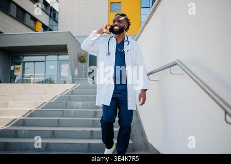 Glücklicher schwarzer Arzt in Kaffeepause, der im Krankenhaus auf dem Handy spricht. Stockfoto