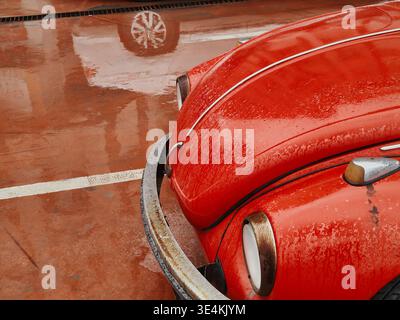 Eine detailreiche Aufnahme der Motorhaube und des Stoßfängers eines klassischen roten Autos, die mit Regentropfen bedeckt sind. Die glänzende nasse Oberfläche des Gehäuses spiegelt das Fahrzeug wider Stockfoto