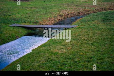 Ein ruhiger Außenfoto einer einfachen Holzplankenbrücke, die einen schmalen, gewundenen Bach überquert. Der Bach fließt durch ein lebhaftes grünes Feld, sein dunkles Wasser Stockfoto