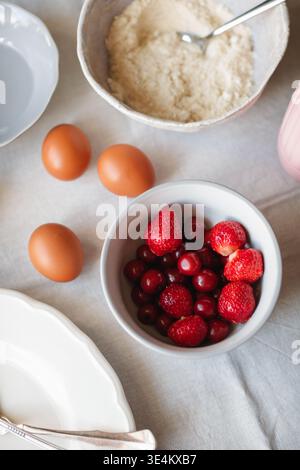 Nahaufnahme der Draufsicht verschiedener Backzutaten für die Zubereitung eines Kuchens, einschließlich Eiern, Erdbeeren, Kirschen und glutenfreiem Mandel- und Kokosmehl Stockfoto