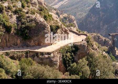 Ein kurviger gepflasterter Pfad führt über steile Felshänge auf dem Montserrat mit Seilbahnanlagen und bewaldeten Klippen im Hintergrund. Stockfoto