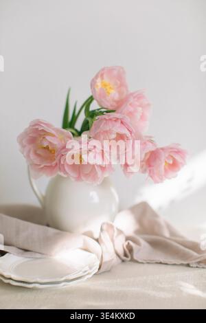 Pinkfarbene Pfingstrosen-Tulpen (Doppel-Tulpen) in einer weißen Kanne auf einem Tisch mit zwei Tellern und einer Leinentischdecke im Sonnenlicht Stockfoto