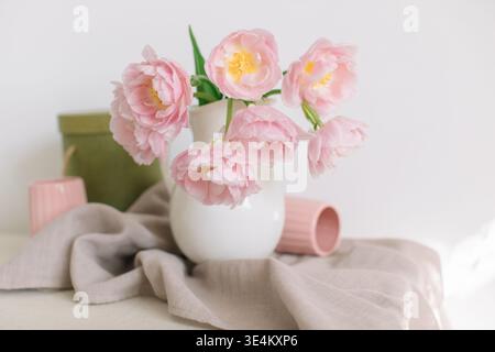 Nahaufnahme von rosa Pfingstrosen-Tulpen (Doppeltulpen) in einem weißen Krug auf einem Tisch mit zwei gerippten Tassen und einer Leinentischdecke Stockfoto