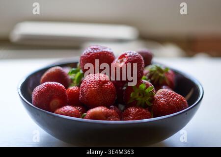 Eine Nahaufnahme von frischen, Reifen roten Erdbeeren mit sichtbaren Wassertropfen auf der Oberfläche, die in einer dunklen Keramikschale gestapelt sind. Die Stockfoto