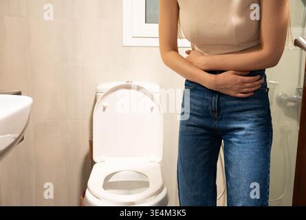 Eine Frau, die sich unwohl in den Magen drückt, steht neben einer Toilette im Badezimmer und vermittelt ein Gefühl von Dringlichkeit und Not. Stockfoto