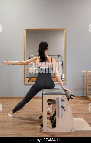 Frau dehnt Seitenkörper auf hölzernem Pilatenkasten, trägt schwarze Sportbekleidung in hellem Studio. Seitliche Stretchpartie, Pilates-Box, Studio, schwarze Sportbekleidung Stockfoto