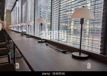 Ein langer Holzschreibtisch verläuft parallel zu einer Wand aus großen Fenstern mit horizontalen Jalousien in einem modernen Büro- oder Kooperationsraum. Eine Reihe von identischen Stockfoto