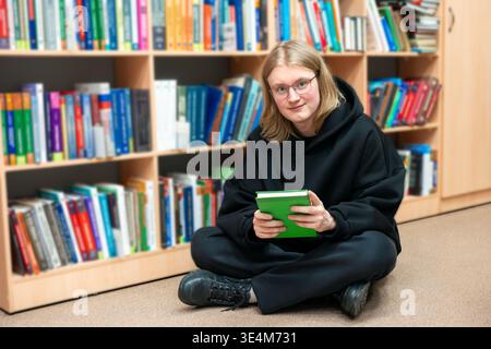 Junger lächelnder Mann, der auf einem Boden in der Bibliothek sitzt und Buch trägt. Kluger Schüler in Brille in der Bibliothek, Bildungskonzept. Regale mit Hintergrund der Buchzeile Stockfoto