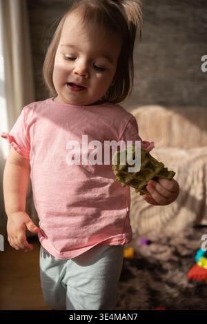 Nahaufnahme eines Kleinkindes, das in warmer Nachmittagssonne in einem gemütlichen Wohnzimmer getaucht ist und ein rosafarbenes Hemd und eine leichte Hose trägt, während er ein grünes hält Stockfoto