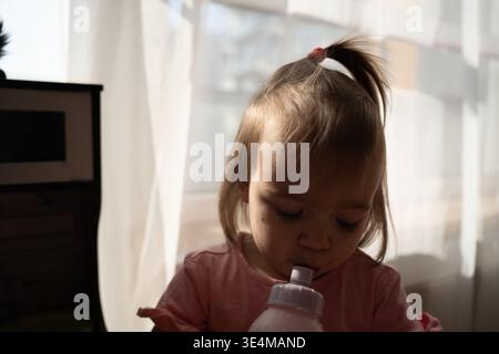Offene Nahaufnahme eines Kleinkindes mit einem winzigen Pferdeschwanz, der aus einer rosa Trinkflasche trinkt, im warmen Morgenlicht durch weiße Vorhänge, weich Stockfoto