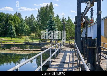 Ein hölzerner Fußgängerweg mit Metallgeländern überquert ruhiges Wasser in einer Industriewasseranlage, wahrscheinlich einem kleinen Wasserkraftwerk. Stockfoto