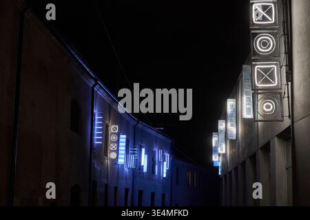 Nächtlicher Blick auf eine enge urbane Gasse zwischen zwei historischen Gebäuden, beleuchtet von zeitgenössischen Lichtkunstinstallationen. Mehrere geometrische Neon Stockfoto