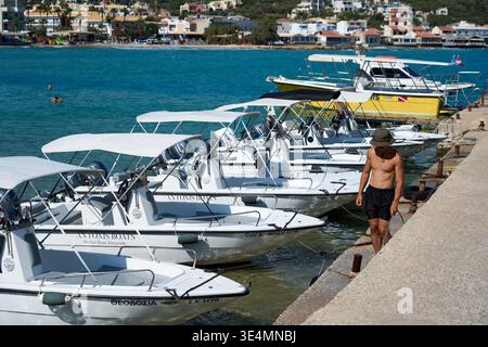 Eine Reihe kleiner Motorboote zum Mieten, die an einem Steinpier in einem Hafen vertäut sind. Ein Mann in Badehosen, der an einem sonnigen Sommertag am Dock entlang läuft. Stockfoto