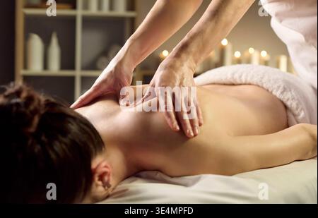 Therapeutin massiert die Schultern der Frau im ruhigen Spa mit warmem Kerzenlicht Stockfoto