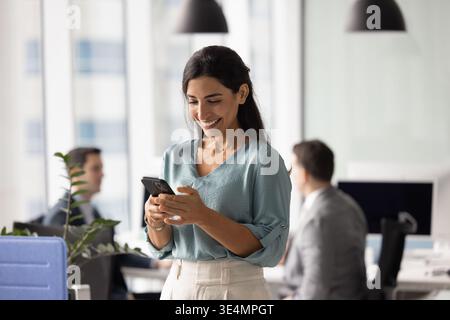 Glückliche, zuversichtliche Geschäftsfrau Latina, die auf einem Smartphone in einem modernen Arbeitsbereich schreibt Stockfoto