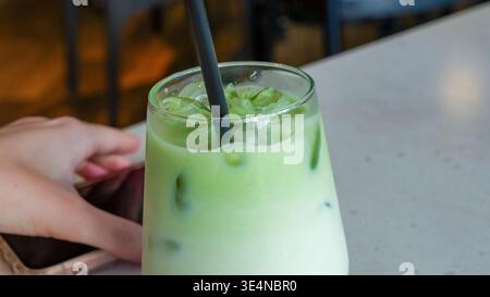 Gefrorener Matcha Latte in einem klaren Glas mit Strohhalm, mit grünen Schichten und Eis, mit einer Hand, die ihn sanft auf einem modernen Tisch hält Stockfoto