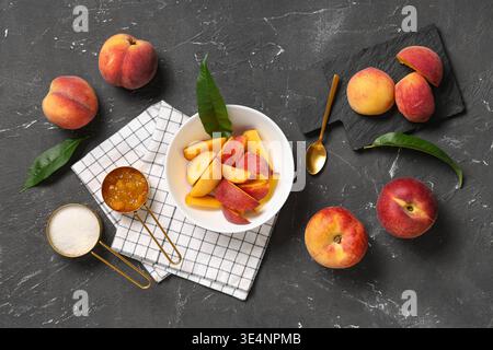 In Scheiben geschnittene Pfirsiche in einer Schüssel, die Tassen Zucker und Marmelade auf grauem Grunge-Hintergrund messen Stockfoto