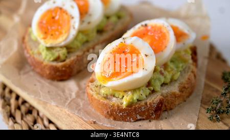 Zwei Scheiben knuspriges Sauerteigbrot mit Avocadopüree und perfekt weich gekochten Eiern mit laufendem Eigelb. Die natürliche Seitenbeleuchtung unterstreicht den Stockfoto