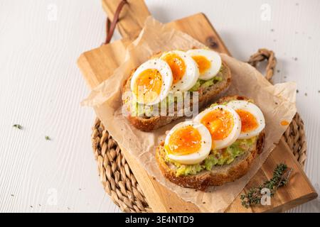 Zwei Scheiben gerösteter Sauerteig, belegt mit Avocadopüree und marmelierten weichen gekochten Eiern. Hochwinkelschuss auf Holzbrett mit Pergamentpapier und Natu Stockfoto