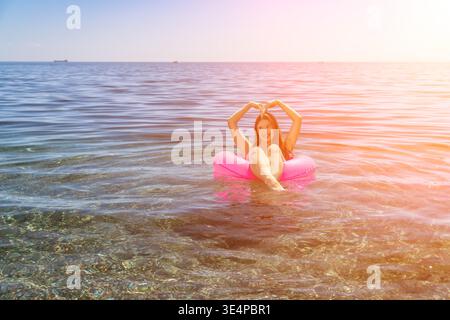Frau, Schwimmen, Urlaub. Glückliche junge Frau schwimmt auf rosa aufblasbarem Ring in klarem Meer und macht Herzform mit Händen während der Sommerferien. Stockfoto