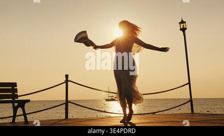 Eine Frau trifft auf See auf den Sonnenaufgang. Emotional drehend mit einem Hut in der Hand auf dem Pier. Traum vom Reisekonzept Stockfoto