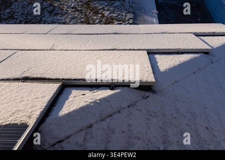 Schneebedeckte Solarpaneele auf einem Dach, beleuchtet durch Sonnenlicht, mit einem frostigen Hinterhof im Hintergrund. Stockfoto