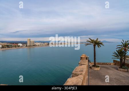 Friedliche Küstenszene. Ruhige Aussicht aufs Meer mit Palmen. Ruhiger Küstenweg mit weit entferntem Stadtbild. Ruhige Strandpromenade mit Palmen Stockfoto