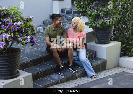 Asiatischer Mann und Afroamerikanerin, die auf Steintreppen sitzen und Hände halten, Pflanzgefäße blühen Stockfoto