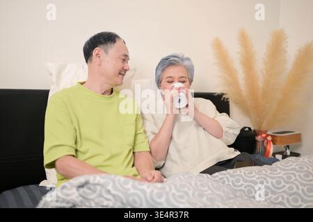 Asiatischer älterer Mann, der seiner Frau ein warmes Getränk gibt, während sie sich im Bett entspannt, und repräsentiert Fürsorge und Zuneigung im häuslichen Alltag || Model veröffentlicht Stockfoto