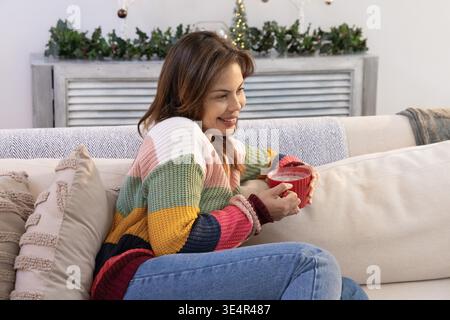 Frau, die auf einem hellen Sofa sitzt und einen gestreiften Pullover trägt, zu Hause einen roten Becher mit einer Kranzgirlande hält. Strickwaren, Damen, Lifestyle, saisonale, Innenausstattung, Ornamente, Grün || Model veröffentlicht Stockfoto