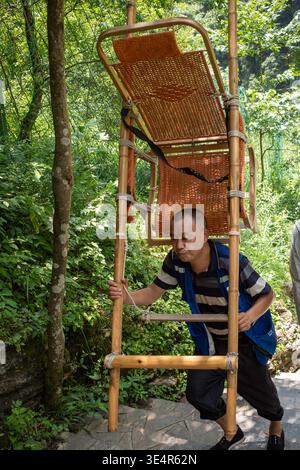 Der Mann trägt Bambusstuhl durch üppig grünen Wald. Natürliches Licht filtert durch dichte Baumkronen. Er greift einen stabilen Holzrahmen und klettert vorsichtig. Stockfoto
