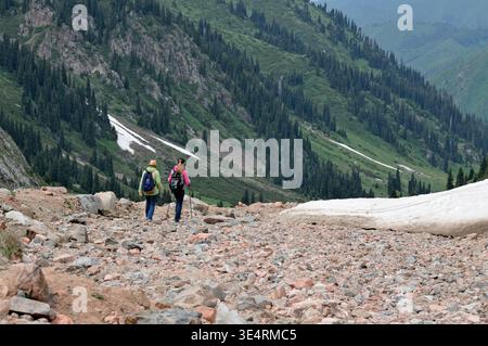 Zwei Wandererinnen mit Rucksäcken, die auf einem felsigen Bergpfad in der Nähe eines Gletschers in der Region Shymbulak spazieren Stockfoto