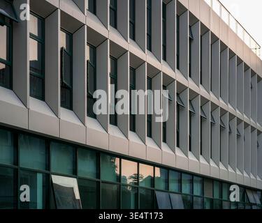Aus nächster Nähe sehen Sie die Fassade eines modernen Bürogebäudes mit sich wiederholenden weißen vertikalen Sonnenschirmen, grauen Paneelen und großen reflektierenden Glasfenstern. Die sc Stockfoto