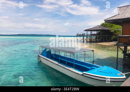 Kleines Motorboot, das neben einer hölzernen Überwasserstruktur auf einer tropischen Insellagune mit klarem türkisfarbenem Wasser und hellem Himmel vor Anker liegt. Stockfoto