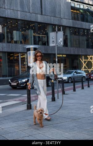 Schöne Frau, die mit ihrem süßen Hund spaziert und einen friedlichen Moment in der Stadt teilt. Lässige Eleganz und starke Bindung an Haustierbesitzer. Stilvolle Frau, die ihre Locken geht Stockfoto