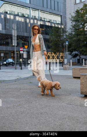Selbstbewusste Frau mit Sonnenbrille geht mit ihrem süßen Hund in die Innenstadt. Helles und legeres Outfit, moderne City-Atmosphäre und Tierleben in einem offenen Stree Stockfoto