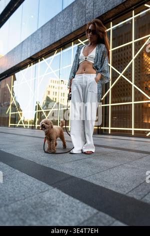 Stilvolle junge Frau, die mit ihrem kleinen Pudelhund in der Stadt spaziert. Moderner urbaner Hintergrund mit Glasreflexionen. Modisches Outfit, selbstbewusste Stockfoto