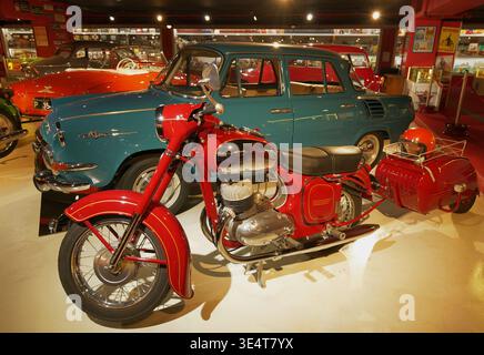 Ein rotes Jawa 350-Motorrad mit einem kleinen Beiwagen ist mit einem Škoda 1000 MB im Hintergrund fotografiert und beleuchtet, um ihre polierten Oberflächen zu zeigen. Stockfoto