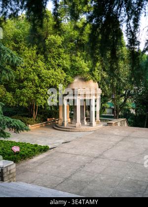 Kleiner weißer kreisförmiger Steinpavillon mit klassischen Säulen, auf einer gepflasterten Plattform, umgeben von üppigen grünen Bäumen in einem öffentlichen Park. Die Szene Stockfoto